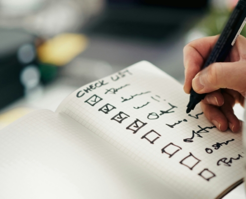 Close-up of a hand writing a checklist in a grid notebook with checked boxes and symbols.