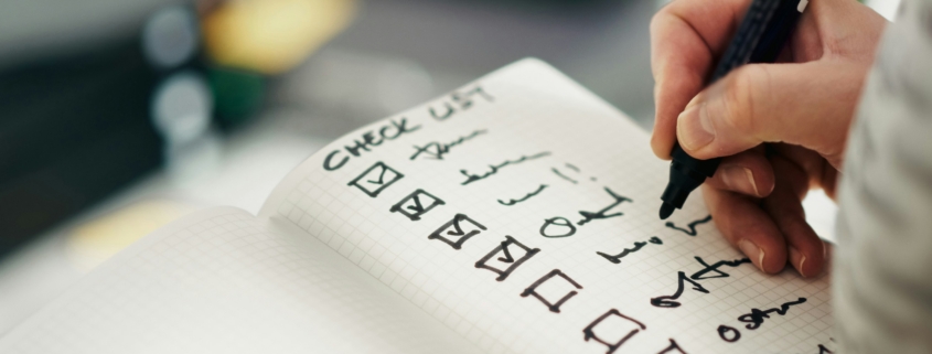 Close-up of a hand writing a checklist in a grid notebook with checked boxes and symbols.