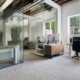 Open-plan office with glass partition walls, exposed ceiling ducts, and a desk area with plants and computer equipment near a large window outside construction materials