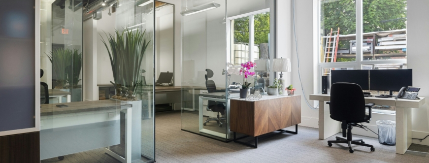 Open-plan office with glass partition walls, exposed ceiling ducts, and a desk area with plants and computer equipment near a large window outside construction materials