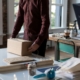 Person in a burgundy shirt lifting or packing a cardboard box at a bright office desk with laptop and photography gear in the background.