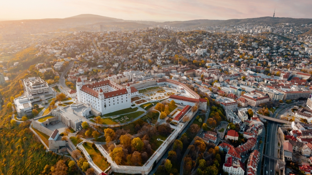 Aerial view of a large white castle with red roofs surrounded by walls and a formal garden, overlooking a densely built city with autumn trees and hills in the distance at sunset.