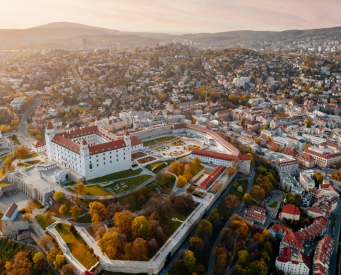 Aerial view of a large white castle with red roofs surrounded by walls and a formal garden, overlooking a densely built city with autumn trees and hills in the distance at sunset.