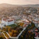 Aerial view of a large white castle with red roofs surrounded by walls and a formal garden, overlooking a densely built city with autumn trees and hills in the distance at sunset.