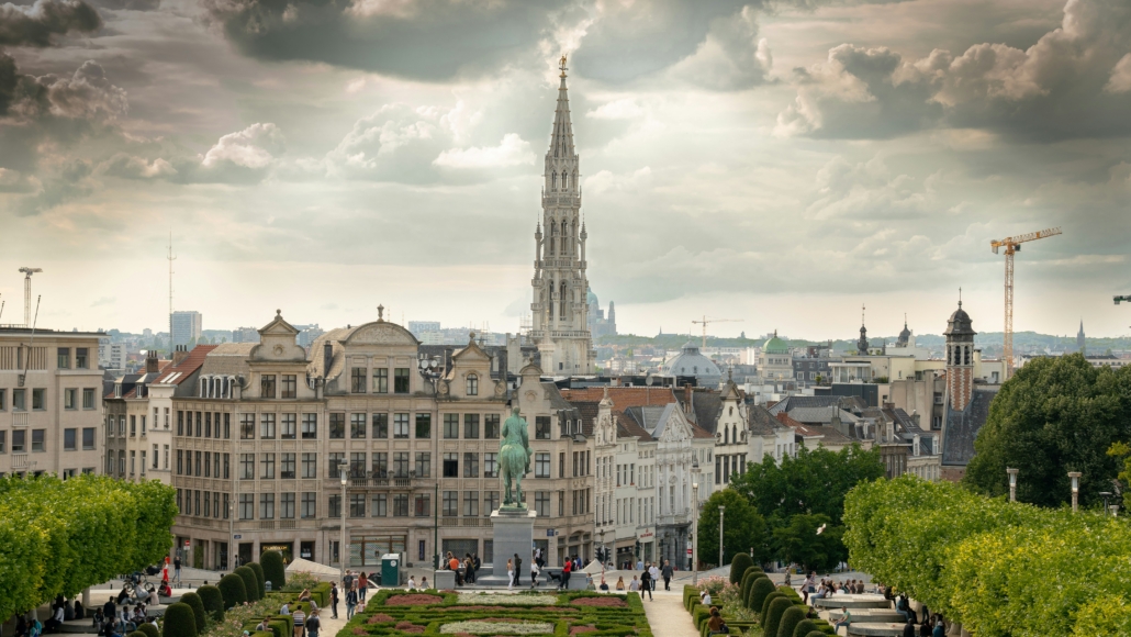 City skyline with a tall Gothic spire rising above beige rooftops and a manicured garden in the foreground under a cloudy sky.
