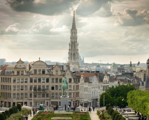 City skyline with a tall Gothic spire rising above beige rooftops and a manicured garden in the foreground under a cloudy sky.