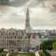 City skyline with a tall Gothic spire rising above beige rooftops and a manicured garden in the foreground under a cloudy sky.