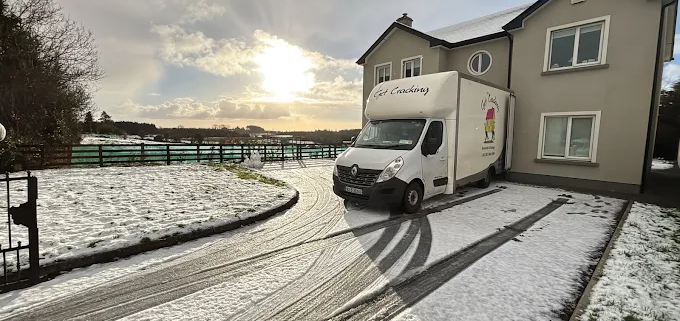 White delivery van parked on a snowy residential driveway beside a two-story beige house, with tire tracks in the snow and a setting sun in the background over a wooden fence.