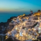 Wide view of Santorini’s whitewashed houses lit at dusk along a cliff, with windmills and the blue Aegean Sea.