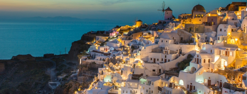 Wide view of Santorini’s whitewashed houses lit at dusk along a cliff, with windmills and the blue Aegean Sea.