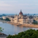 Budapest Parliament Building with red dome along the Danube River, cityscape on the riverbank and a bridge in the foreground, daytime light.