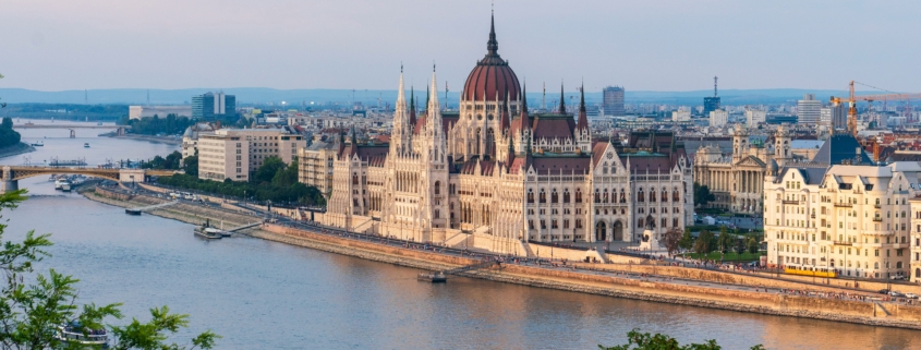 Budapest Parliament Building with red dome along the Danube River, cityscape on the riverbank and a bridge in the foreground, daytime light.