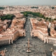 Aerial view of St. Peter's Square in Vatican City, showing the central obelisk, surrounding colonnades, and a crowded plaza below.