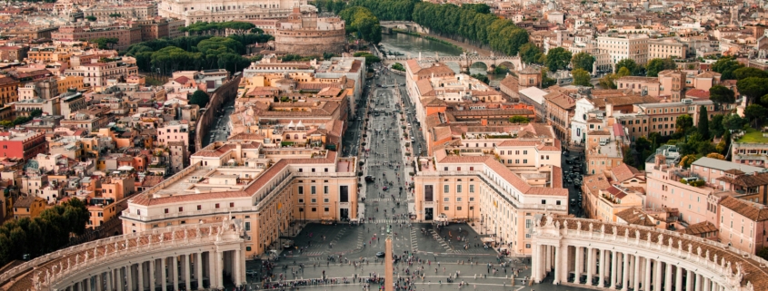 Aerial view of St. Peter's Square in Vatican City, showing the central obelisk, surrounding colonnades, and a crowded plaza below.