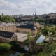 Panoramic view of a historic European town with beige buildings, a river with arches, and church spires atop a hillside under a cloudy sky
