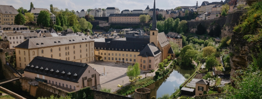 Panoramic view of a historic European town with beige buildings, a river with arches, and church spires atop a hillside under a cloudy sky