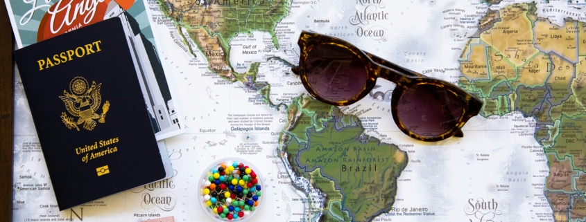 Passport and travel brochures rest on a world map with a pair of sunglasses and colorful pins for planning a trip.