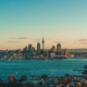 City skyline along a harbor at sunset, with cranes left and tall buildings near the center under a blue-pale sky.