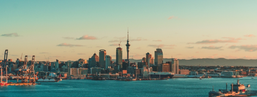 City skyline along a harbor at sunset, with cranes left and tall buildings near the center under a blue-pale sky.