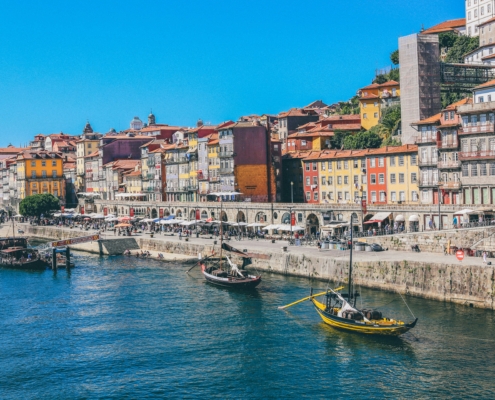 Colorful row houses line a riverside harbor with small boats and a bright blue sky above.