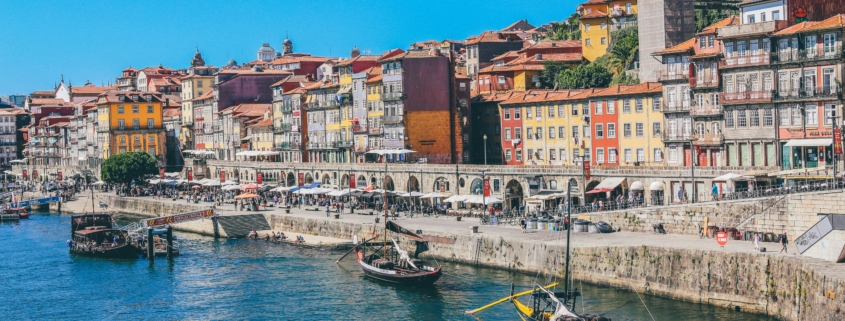 Colorful row houses line a riverside harbor with small boats and a bright blue sky above.
