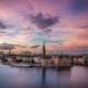 City skyline at sunset with a tall church spire rising above historic buildings along the river.