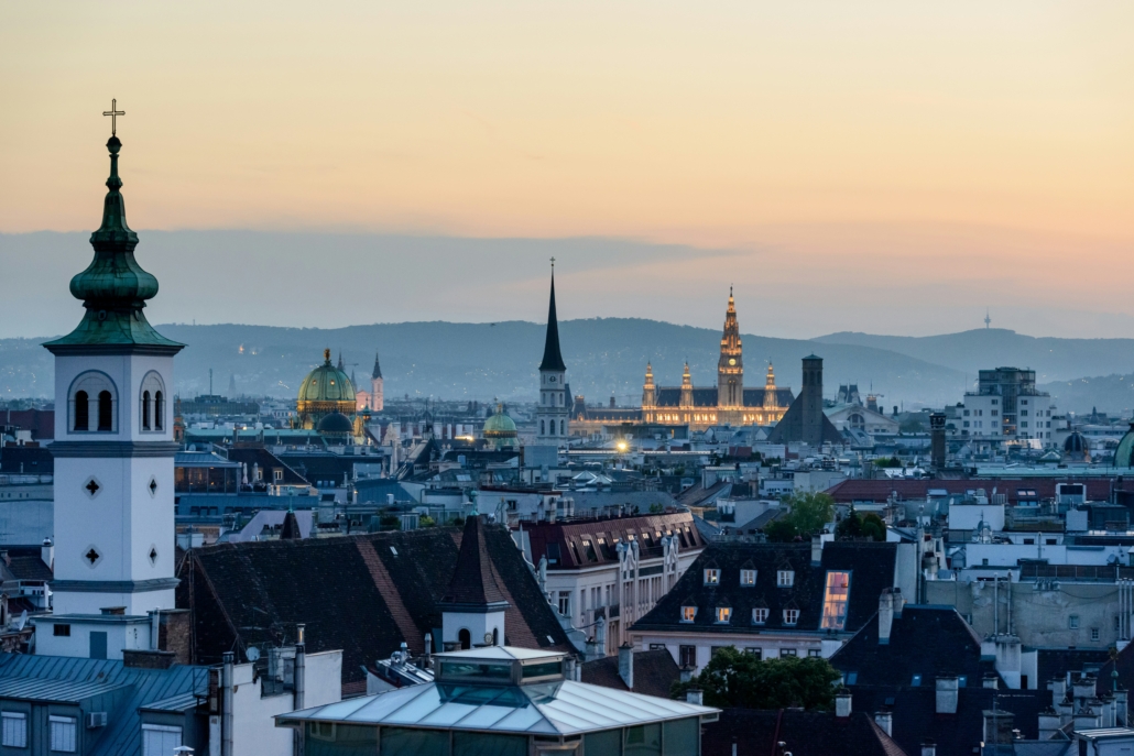 Sunset cityscape with church spires and green-domed roof over a dense rooftop skyline.