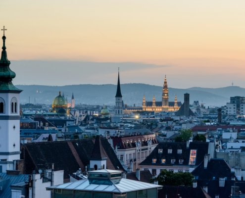 Sunset cityscape with church spires and green-domed roof over a dense rooftop skyline.