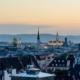 Sunset cityscape with church spires and green-domed roof over a dense rooftop skyline.