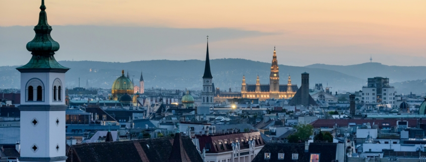Sunset cityscape with church spires and green-domed roof over a dense rooftop skyline.